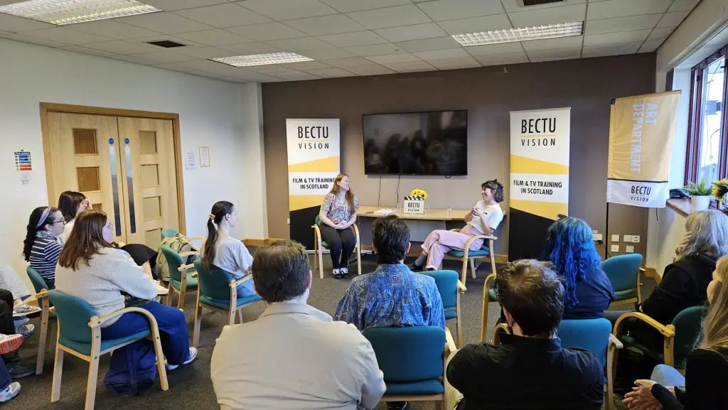 BECTU Vision's New Entrants' Base Drop-in session in a meeting room with BECTU Vision branded banners each side of Jena Hunter, Learning Co-ordinator from BECTU Vision and Jodi Webster Art Department practitioner in Q&A session with audience listening