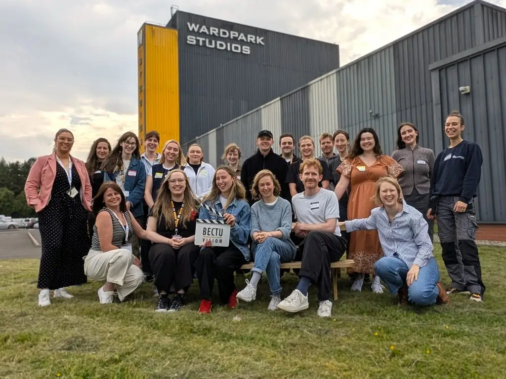 A group of 21 people smiling for a photo in front of Wardpark Studios, holding a BECTU Vision clapperboard